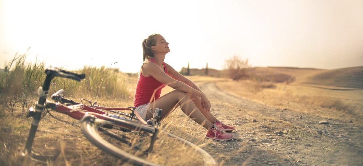 Full body of female in shorts and top sitting on roadside in rural field with bicycle near and enjoying fresh air with eyes closed