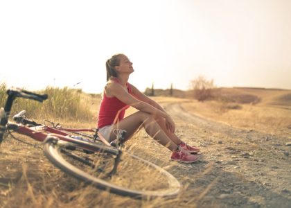 Full body of female in shorts and top sitting on roadside in rural field with bicycle near and enjoying fresh air with eyes closed